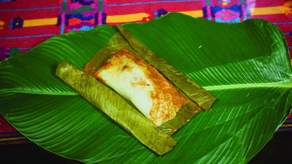 Receta tamales de arroz guatemaltecos: Conozca este manjar del altiplano – SoyMigrante.com REVISTA Pan de muerto tradicional en hoja de plátano, símbolo de la cultura y festividad mexicana, presentado en una celebración popular con colores vibrantes de fondo. – SoyMigrante.com REVISTA