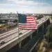 Bandera de EE.UU. ondeando en una autopista con ciudad en el fondo, símbolo de migración y cultura estadounidense, imagen representativa para la revista SoyMigrante.com REVISTA.