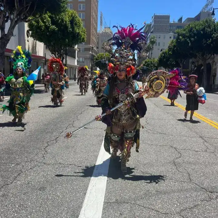Arlequín en carnaval con vestuario colorido y elaborado, participando en desfile callejero con otros bailarines y músicos, celebrando cultura latinoamericana en ciudad moderna, en un día soleado. – SoyMigrante.com REVISTA