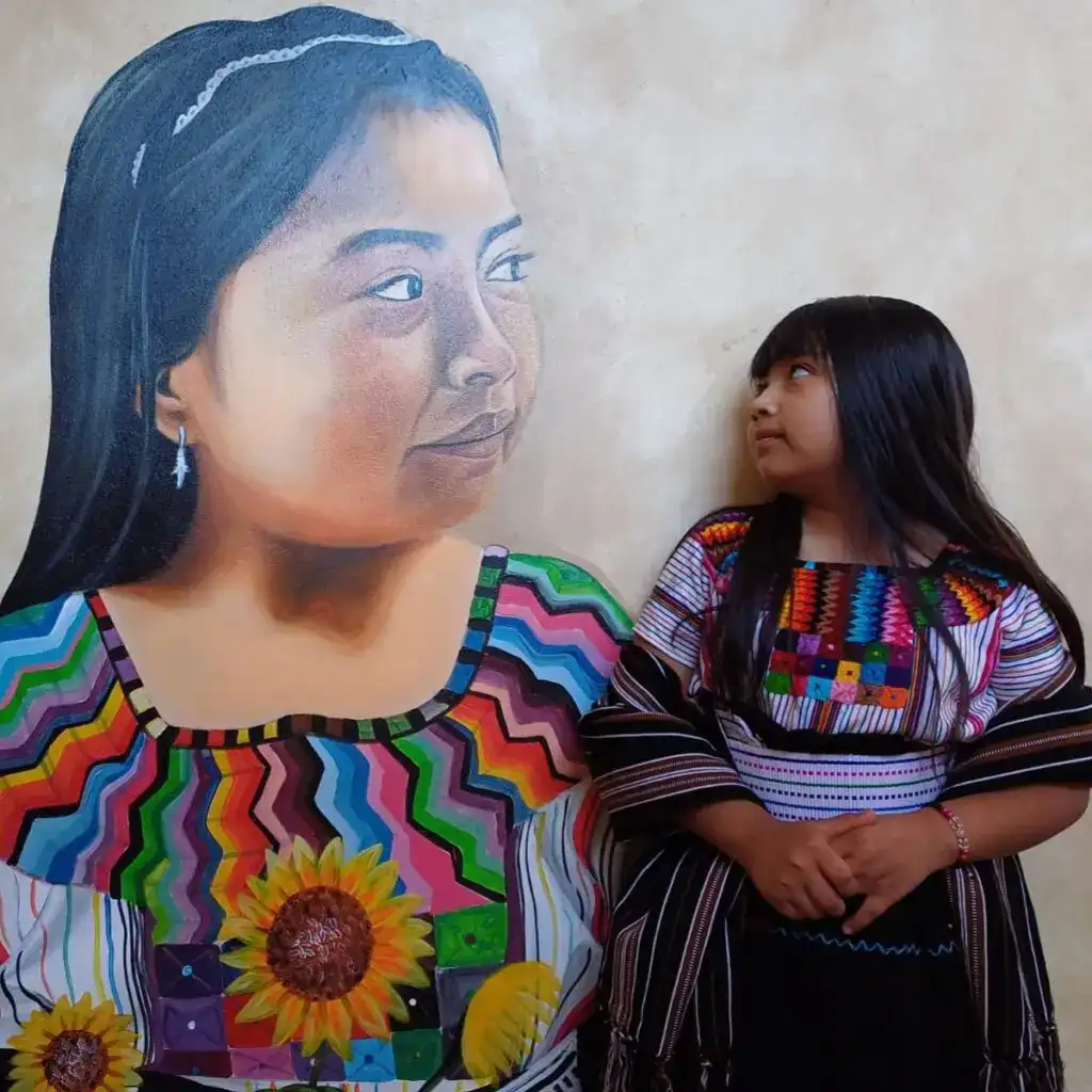 Colorida pintura mural de una mujer indígena vestida con ropa tradicional mexicana, acompañada por una niña en una exposición cultural, representando la diversidad y cultura del país. – SoyMigrante.com REVISTA