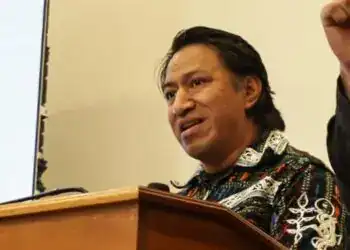 Voz enérgica de un conferencista latinoamericano en una reunión o seminario, destacando la participación cultural y educativa en la revista SoyMigrante.com REVISTA.