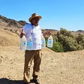 Hombre en un paisaje árido sosteniendo botellas de agua, símbolo de resistencia y esfuerzo en trabajos migrantes en entornos rurales. – SoyMigrante.com REVISTA