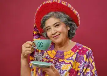 Disfruta del arte y la cultura con una mujer mayor vestida con ropa tradicional mexicana y sombrero colorido, sosteniendo una taza de café en un fondo rojo. – SoyMigrante.com REVISTA
