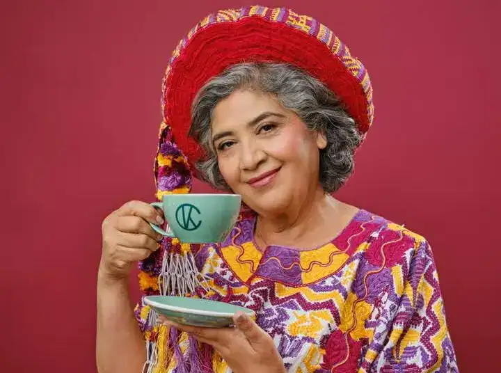 Disfruta del arte y la cultura con una mujer mayor vestida con ropa tradicional mexicana y sombrero colorido, sosteniendo una taza de café en un fondo rojo.