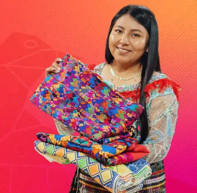 Tejedora indígena con textiles coloridos y tradicionales, mostrando su cultura y arte en un fondo vibrante en la revista SoyMigrante.com. – SoyMigrante.com REVISTA