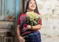 Her royalty-free photo of a smiling woman holding a bouquet of flowers, wearing traditional indigenous clothing, standing against a rustic wall in a rural setting, representing migrant culture and heritage. – SoyMigrante.com REVISTA