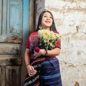 Her royalty-free photo of a smiling woman holding a bouquet of flowers, wearing traditional indigenous clothing, standing against a rustic wall in a rural setting, representing migrant culture and heritage.