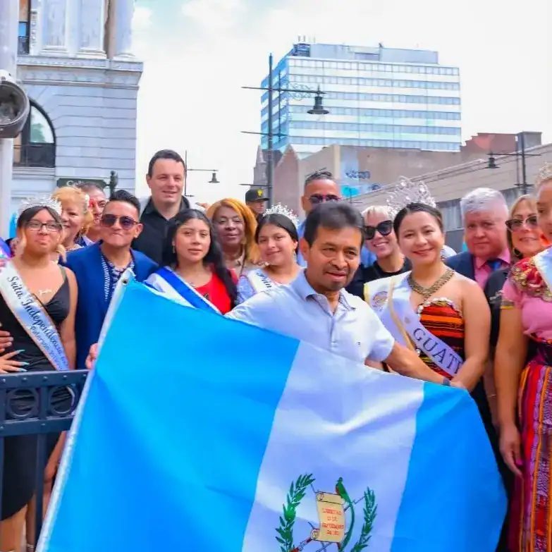 Rostro de un grupo diverso de personas en una celebración en la calle, algunas con coronas y banderas, en un evento que destaca la cultura y comunidad migrante guatemalteca en un entorno urbano.