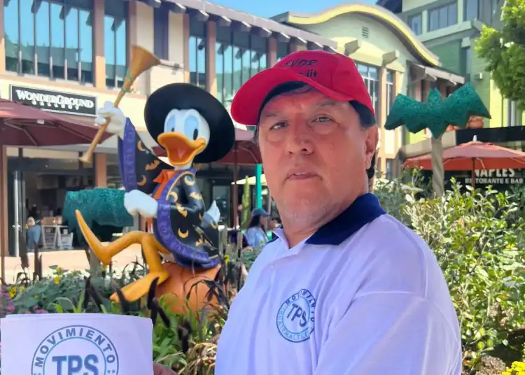 Turista en un centro comercial de EE.UU., sostiene una hoja con el logotipo de TPS, en un entorno urbano con decoraciones de Halloween y figura de Mickey Mouse disfrazada.