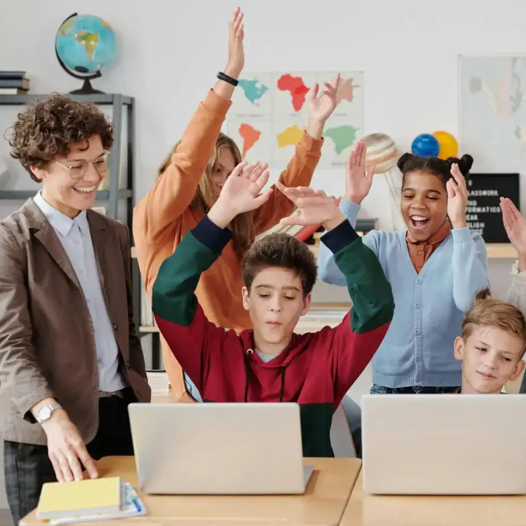 Estudiantes y profesora en un aula de clases, usando laptops, celebrando algo juntos, entorno educativo y multicultural en escuela moderna. – SoyMigrante.com REVISTA