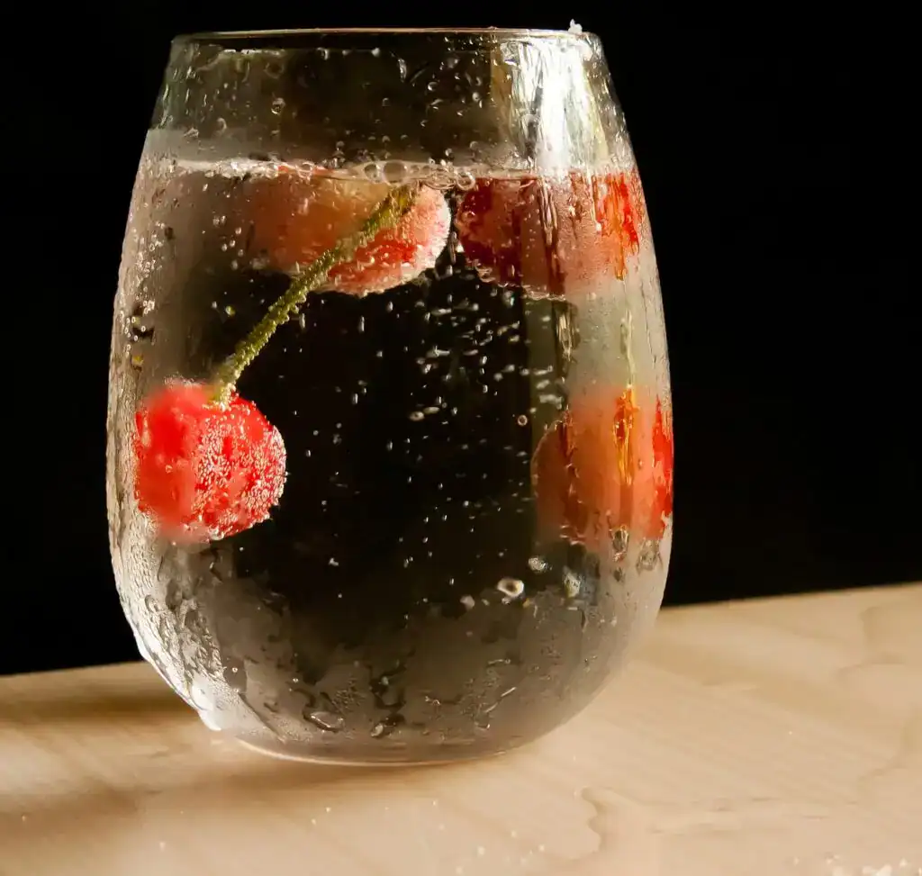 Refresco con cerezas y burbujas, en un vaso transparente sobre fondo negro, resaltando la frescura y la efervescencia ideal para bebidas de verano. – SoyMigrante.com REVISTA