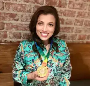 Medalla de oro en igualdad y migración, mujer sonriendo con vestido colorido y fondo de ladrillos en revista SoyMigrante.com REVISTA. – SoyMigrante.com REVISTA