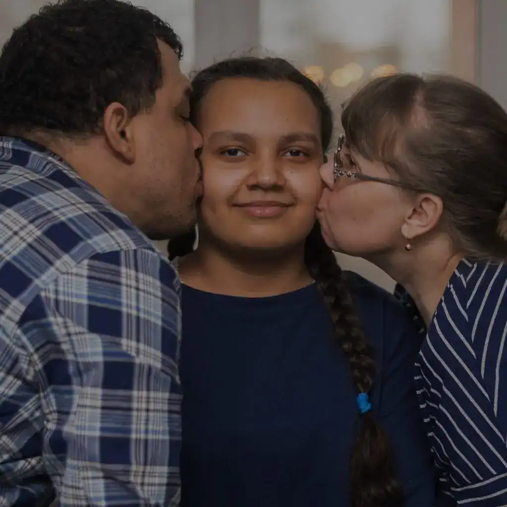 - Persona inmigrante rodeada de madre y padre dándole un beso en las mejillas, mostrando amor y apoyo en su proceso migratorio.