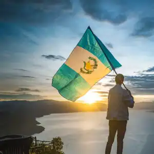 Bandera de Guatemala ondeada por persona en paisaje al atardecer, símbolo cultural y de identidad.
