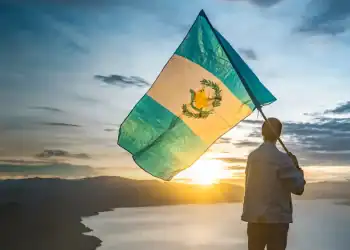 Bandera de Guatemala ondeada por persona en paisaje al atardecer, símbolo cultural y de identidad.