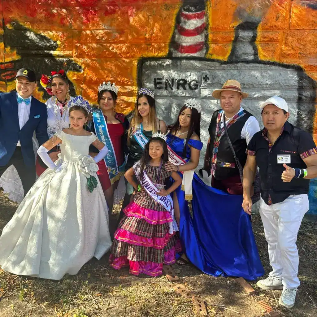 Disfruta de la diversidad cultural en la celebración con personas en trajes tradicionales frente a un mural que menciona la generación de energía sostenible, promoviendo el orgullo y la identidad de la cultura latina. – SoyMigrante.com REVISTA