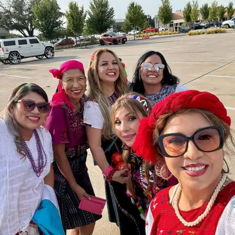 Activistas migrantes con atuendos tradicionales y sonrisas en un evento al aire libre, promoviendo la cultura y la integración en la comunidad en un día soleado en el estacionamiento de una ciudad laboral. – SoyMigrante.com REVISTA