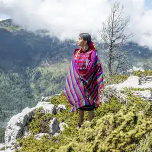 Colorida mujer indígena con traje tradicional en la montaña, resaltando la cultura migrante y la diversidad en América Latina.
