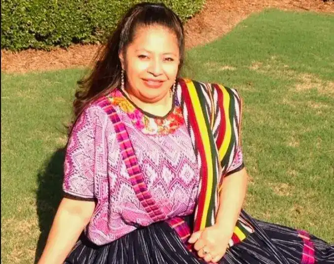 Retrato de una mujer migrante con vestimenta tradicional, sonriendo y sentada en un jardín.
