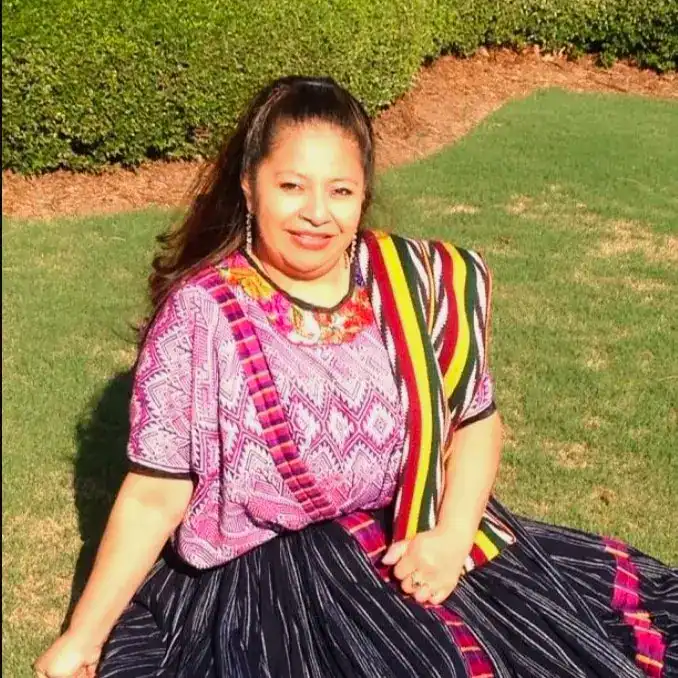 Miriam Tepe migrante guatemalteca en Oklahoma – SoyMigrante.com REVISTA Retrato de una mujer migrante con vestimenta tradicional, sonriendo y sentada en un jardín.