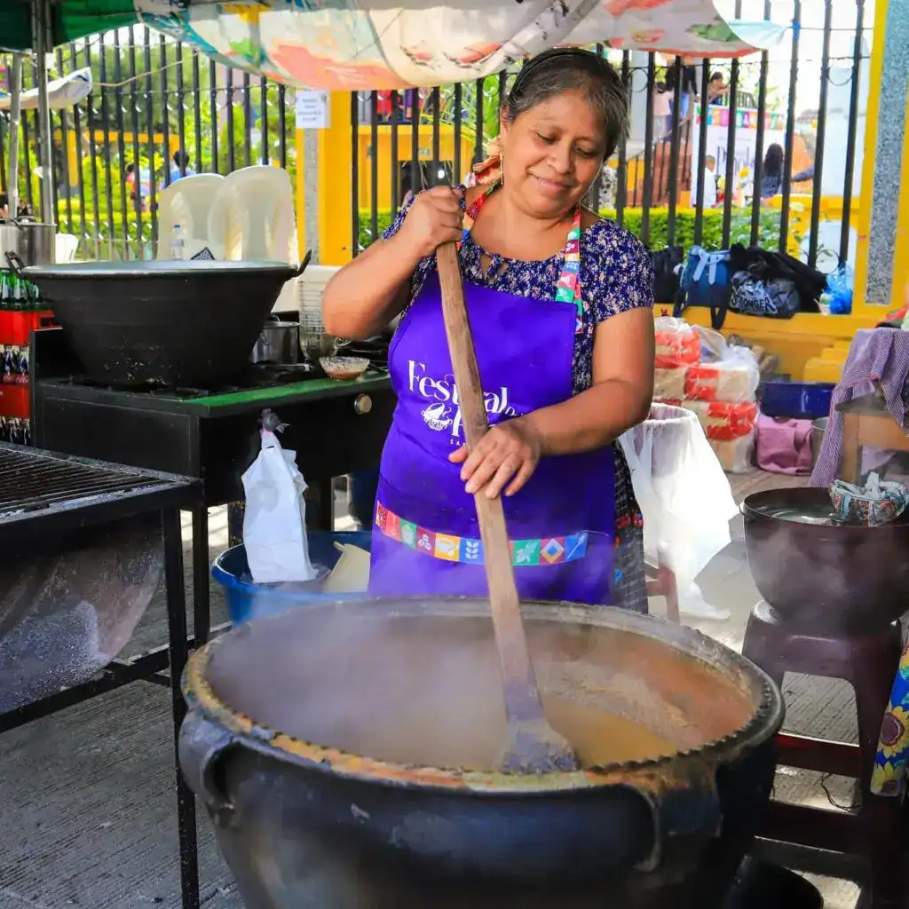 Artesana preparando comida en una feria cultural y gastronómica en un puesto callejero en un ambiente festivo y vibrante en México.