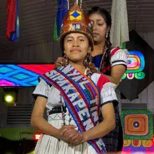 Orgullosa mujer indígena en vestimenta tradicional con banda que dice "Soy Migrante" en evento cultural.
