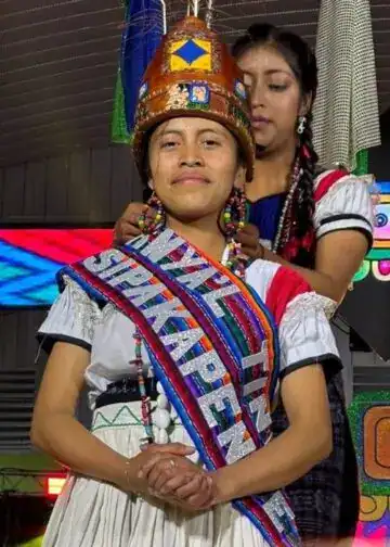 Reina del pueblo sipakapense, Credito Ixchel Producciones. – SoyMigrante.com REVISTA Orgullosa mujer indígena en vestimenta tradicional con banda que dice "Soy Migrante" en evento cultural. – SoyMigrante.com REVISTA