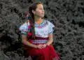 Mujer indígena con vestimenta tradicional sentada entre lava volcánica, símbolo de resistencia y cultura en México, en una imagen que refleja identidad y raíces. – SoyMigrante.com REVISTA