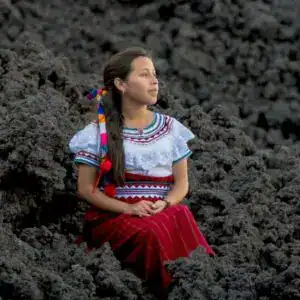 Mujer indígena con vestimenta tradicional sentada entre lava volcánica, símbolo de resistencia y cultura en México, en una imagen que refleja identidad y raíces.