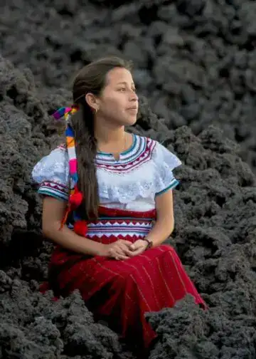 Comunidad Q'anjob'al vive en - San Pedro Soloma - San Juan Ixcoy - Santa Cruz Barillas - Santa Eulalia. Foto Abel Juarez – SoyMigrante.com REVISTA Mujer indígena con vestimenta tradicional sentada entre lava volcánica, símbolo de resistencia y cultura en México, en una imagen que refleja identidad y raíces. – SoyMigrante.com REVISTA