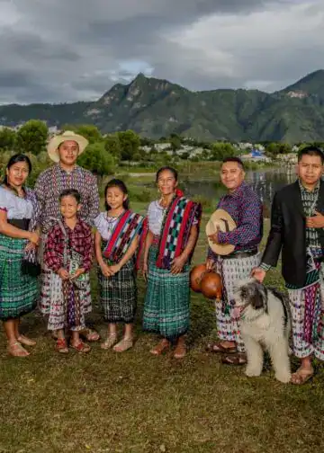 El artista Tz'utujil Juan Bautista Navichoc comparte esta fotografía de su archivo personal junto a su familia, tomada en San Pedro La Laguna, y al fondo se aprecia el Lago de Atitlán. Retrato tomado por Carlos Francisco Pop – SoyMigrante.com REVISTA Gente indígena con trajes tradicionales y paisaje montañoso al fondo, reflejando cultura y tradición en México. – SoyMigrante.com REVISTA