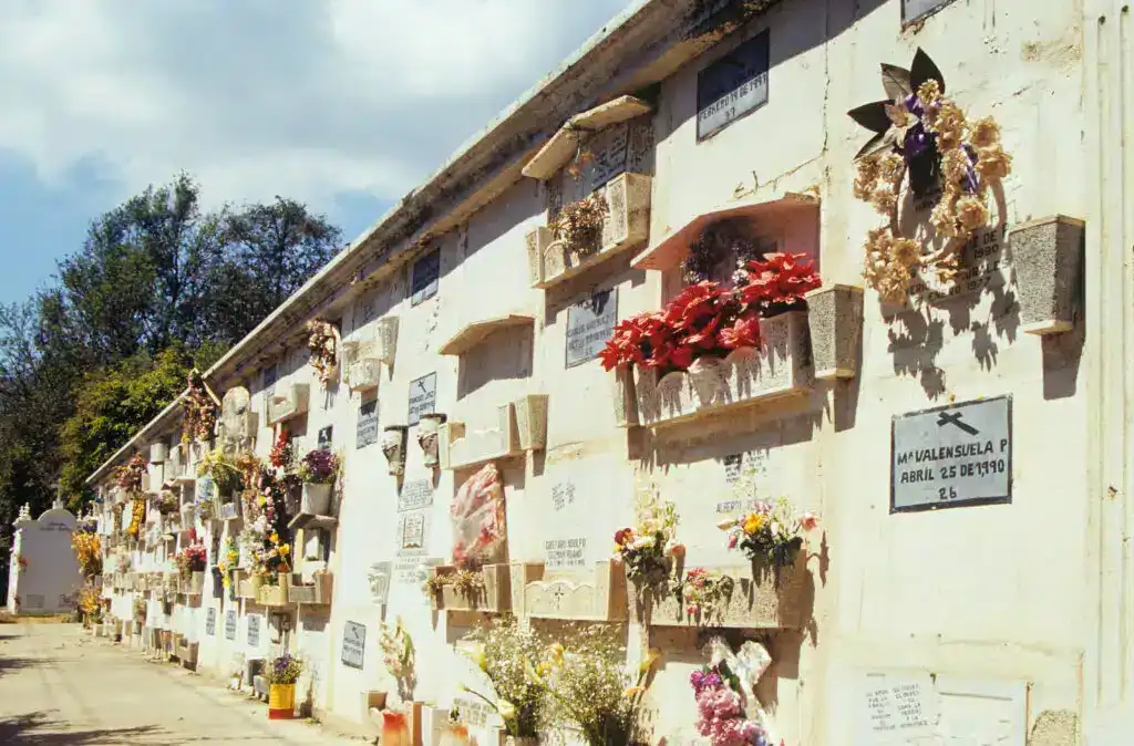 Florería y monumento en un cementerio mexicano con nichos y placas conmemorativas, decoradas con flores frescas y artificiales, en un día soleado, símbolo de memoria y homenaje. – SoyMigrante.com REVISTA