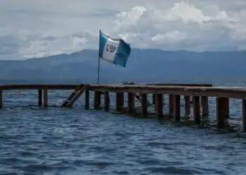 Bandera de Guatemala en un muelle sobre el agua, con montañas al fondo y cielo nublado, representando la migración y presencia de guatemaltecos en el extranjero.