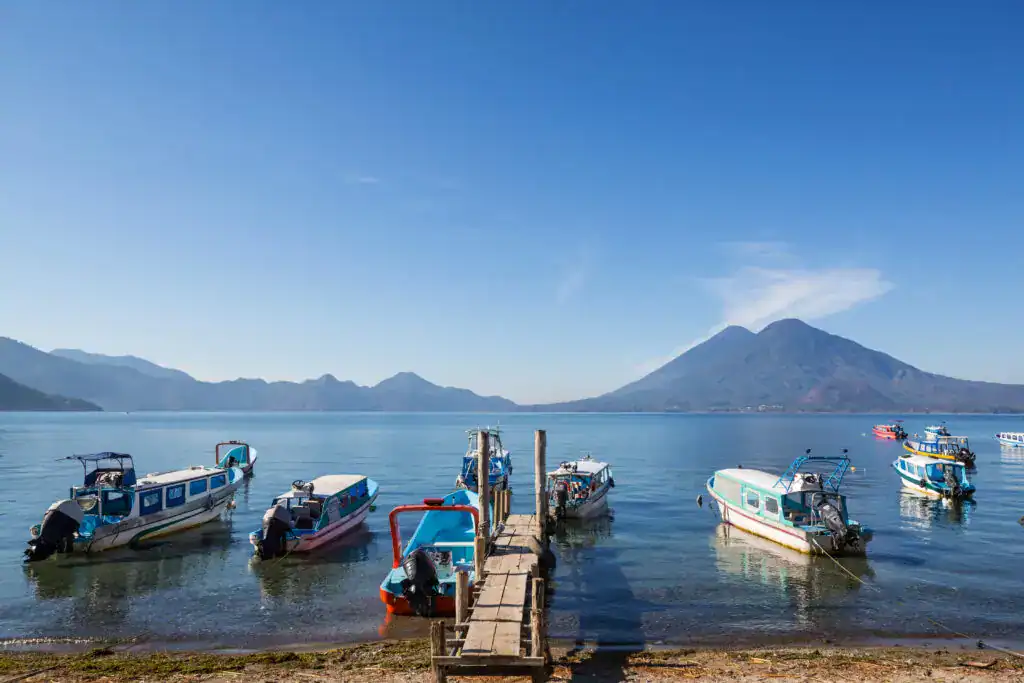 Embarcaciones en el lago con volcán en el fondo, paisaje natural, turismo en Guatemala, vista del Volcán de Fuego, instrumentos náuticos en barco, clima despejado, destino turístico en Centroamérica. – SoyMigrante.com REVISTA