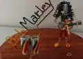 Creador de arte con latas recicladas de colores y musicalina que forma a un personaje con guitarra y nombre Marely, promoviendo la creatividad y el reciclaje en la comunidad migrante.