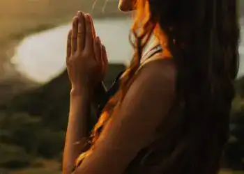 Piel de una mujer con cabello rizado en posición de oración frente a un atardecer, simbolizando paz, esperanza y reflexión, en un entorno natural junto a un río.