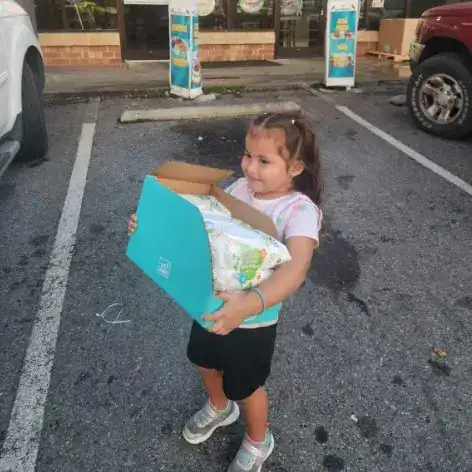 donativos morganton – SoyMigrante.com REVISTA Joven niña con caja de pañales en parque de estacionamiento, invitando a la reflexión sobre derechos y bienestar infantil en comunidades migrantes. – SoyMigrante.com REVISTA