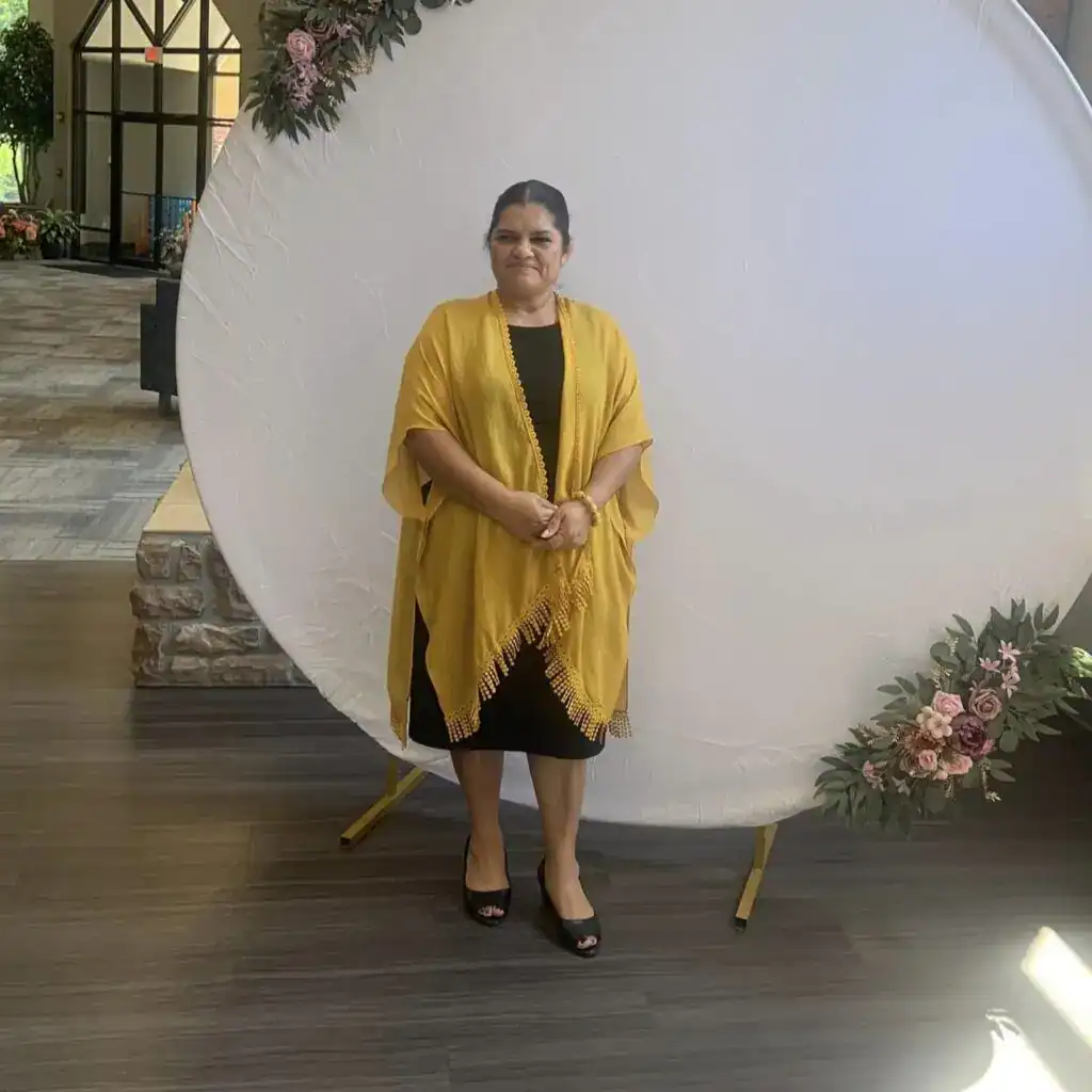 Celebración cultural y migrante en evento, mujer con vestimenta amarilla en fondo decorativo floral, destacando historias de migrantes, comunidad, cultura y solidaridades en revista SoyMigrante.comRevista.