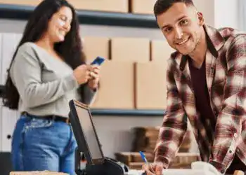 Trabajador migrante en oficina, posando sonriendo y firmando documento, entorno de trabajo con computadora y cajas de archivo, representando historias de migrantes y trabajo en el extranjero en SoyMigrante.com REVISTA.