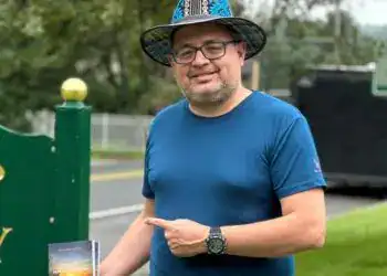 Discurso de migrante latinoamericano en parque público, promoviendo integración y cultura, con libro en mano, vestido con camiseta azul y sombrero colorido.