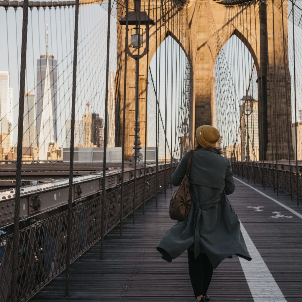 Puente de Brooklyn con mujer caminando, símbolo de inmigración y diversidad en Nueva York, ideal para contenidos relacionados con migrantes, cultura y estilo de vida en Estados Unidos. – SoyMigrante.com REVISTA