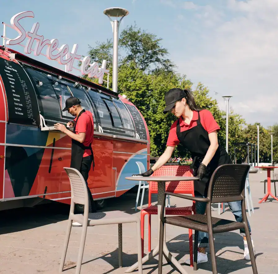migrantes remesas – SoyMigrante.com REVISTA Street food food truck en la calle, con camareras sirviendo bebidas y pagando en la caja, en un día soleado y con árboles verdes al fondo, muestra el crecimiento del turismo y la gastronomía callejera en ciudades modernas.