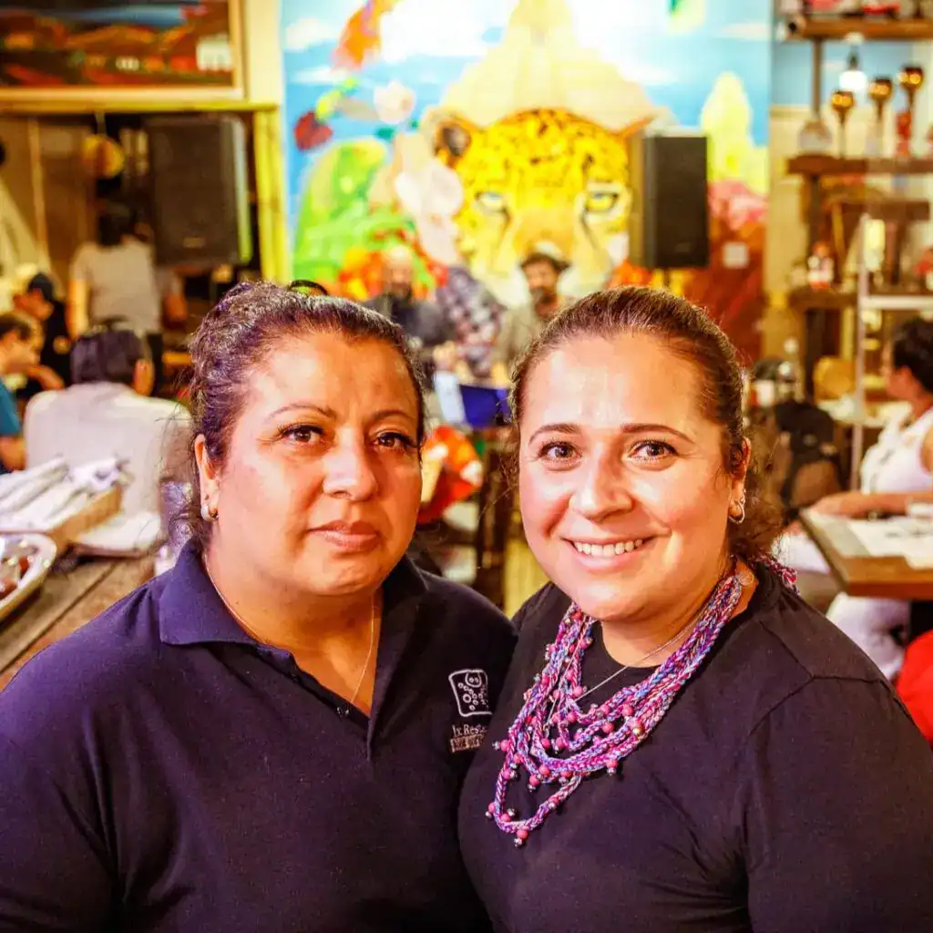 Trabajadoras migrantes en un restaurante latino con decoración colorida y mural de jaguar en el fondo, mostrando la cultura y comunidad migrante en eventos culturales y sociales. – SoyMigrante.com REVISTA