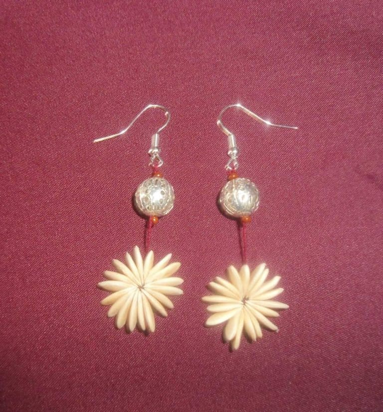 Juego de aretes artesanales con bolas de plata y flores blancas en un fondo rosa profundo.