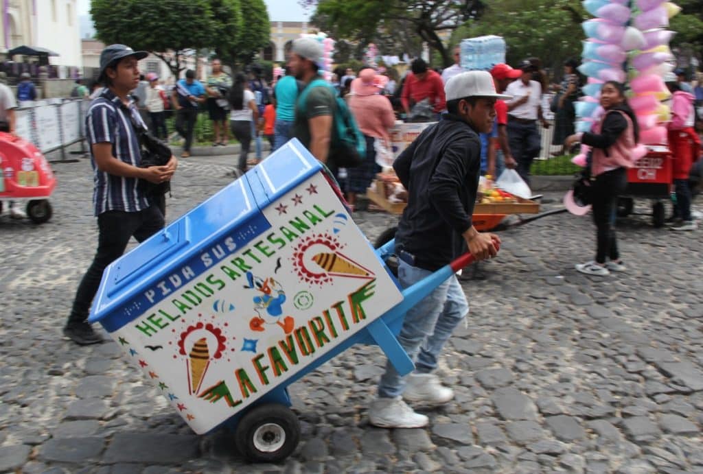 Heladero ambulante en festivales mexicanos, promoviendo helados artesanales y tradicionales en calles de ciudad. Personas disfrutando de la feria y ambiente festivo en un día soleado. – SoyMigrante.com REVISTA