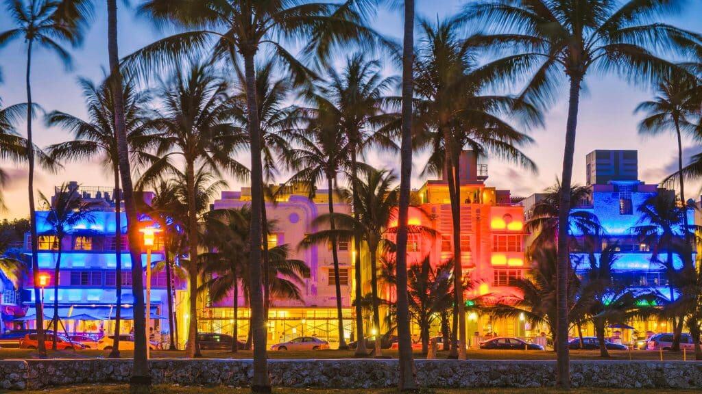 Vibrante imagen de Miami con edificios iluminados en colores neón y muchas palmeras, reflejando la cultura, migrantes y estilo de vida en ciudades cosmopolitas del sur de Florida.