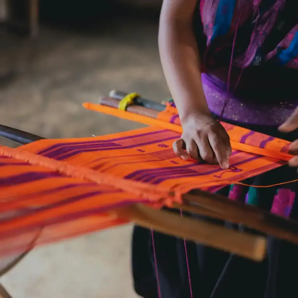 Tela de trabajo artesanal con tejido colorido y patrón tradicional, realizada por una mujer indígena en un entorno rural de Latinoamérica. – SoyMigrante.com REVISTA