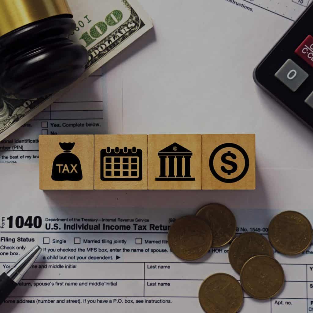 Dinero en efectivo, lápiz y bloques con íconos de impuestos, calendario, banco y dinero, relacionados con la fiscalidad para migrantes y la declaración de impuestos en EE. UU. – SoyMigrante.com REVISTA