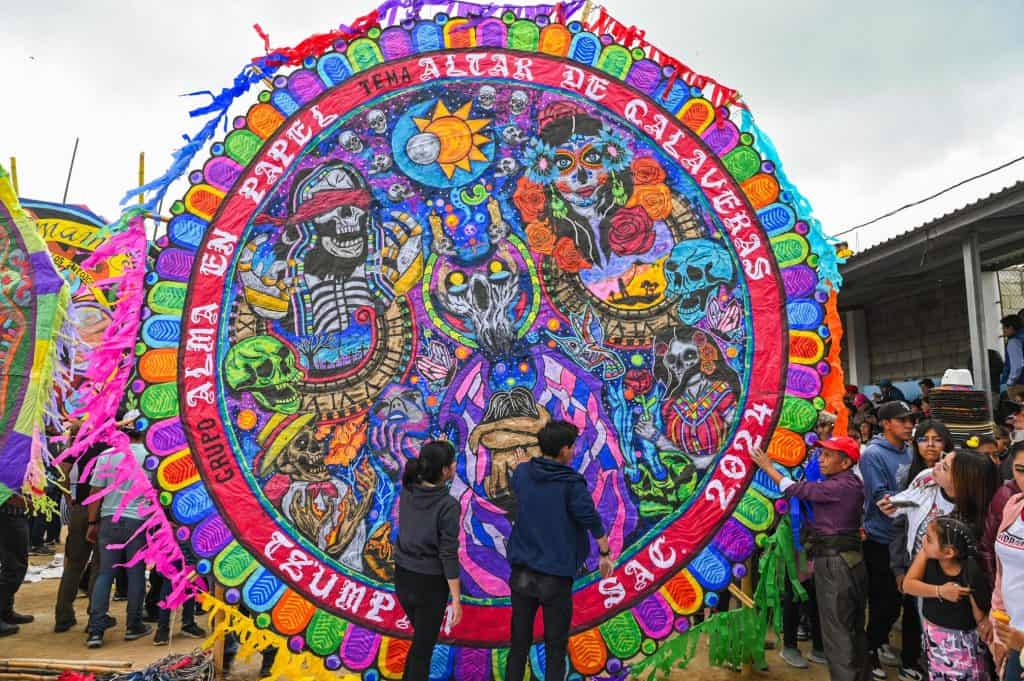 Colorido altar de calaveras en una celebración tradicional con personas observando y participando, representando la cultura mexicana del Día de Muertos y sus expresiones artísticas y religiosas. – SoyMigrante.com REVISTA