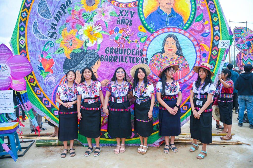 Colorido desfile de mujeres en traje tradicional con un enorme escolta de paracaídas, celebrando 25 años de orgullo y barriletes en la festividad de Oaxaqueña en las Barrileteras. – SoyMigrante.com REVISTA